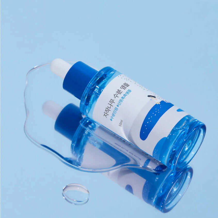 Bottle of ROUND LAB Birch Moisture Ampoule 50ml lying on a blue surface with droplets.
