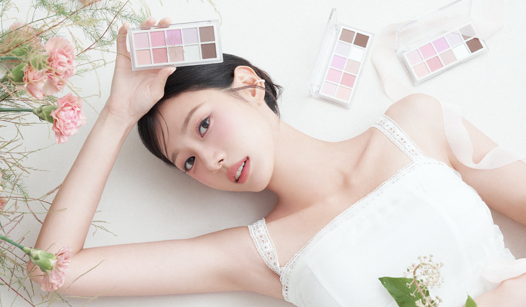 Woman lying down surrounded by pink and brown eyeshadow palettes and flowers on a soft white background.