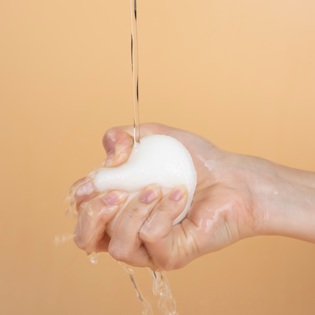 Hand squeezing a foam ball with water dripping against a peach background, illustrating cleansing product application.