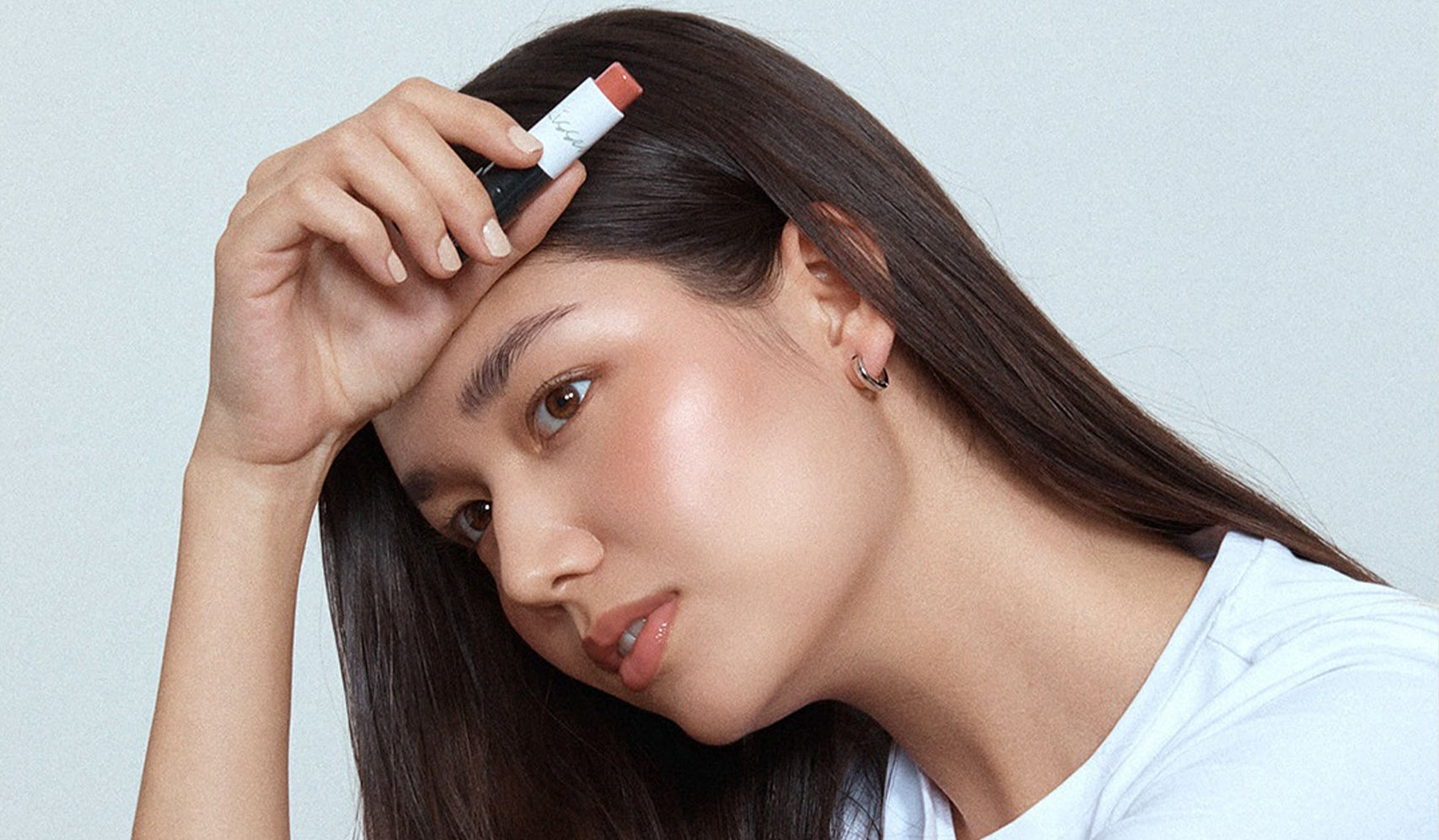 Woman holding a lipstick near her forehead wearing a white t-shirt, showcasing vibrant makeup.