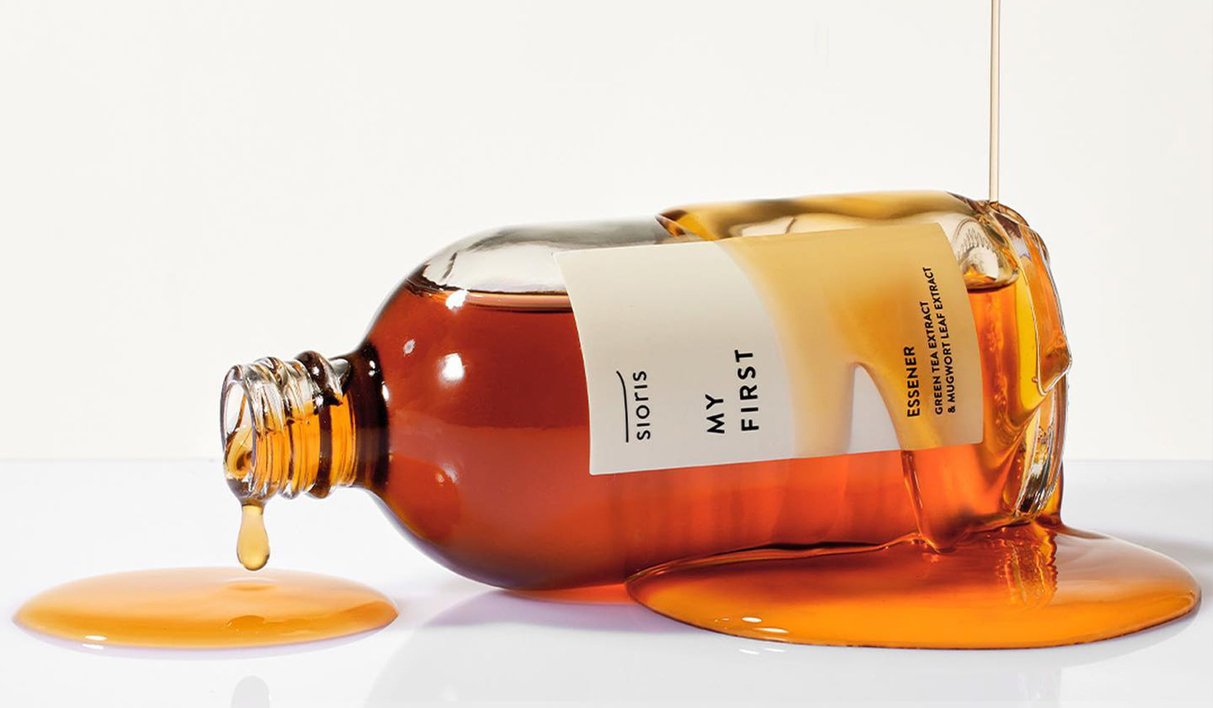 Spilled bottle of essence with liquid flowing, label reading 'My First' on a white background, showcasing packaging and product texture.