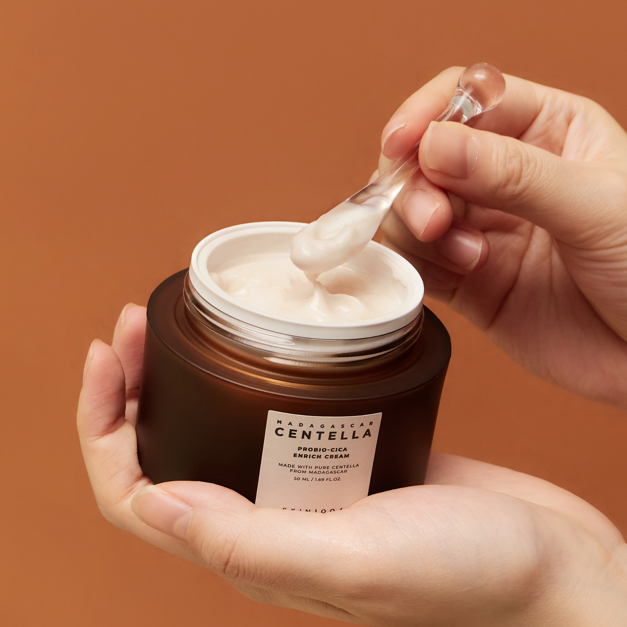 A jar of SKIN1004 Madagascar Centella Probio-cica Enrich Cream 50ml being applied with a spatula against a brown background.