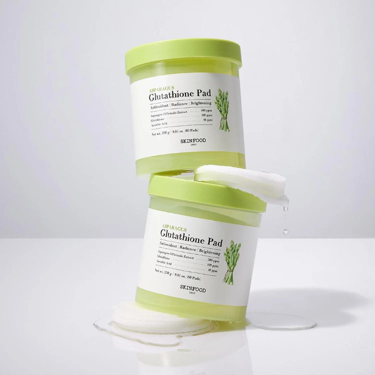Stacked containers of SKINFOOD Asparagus Glutathione Pad (60 Pads) with pads and moisture visible, emphasizing skincare benefits.