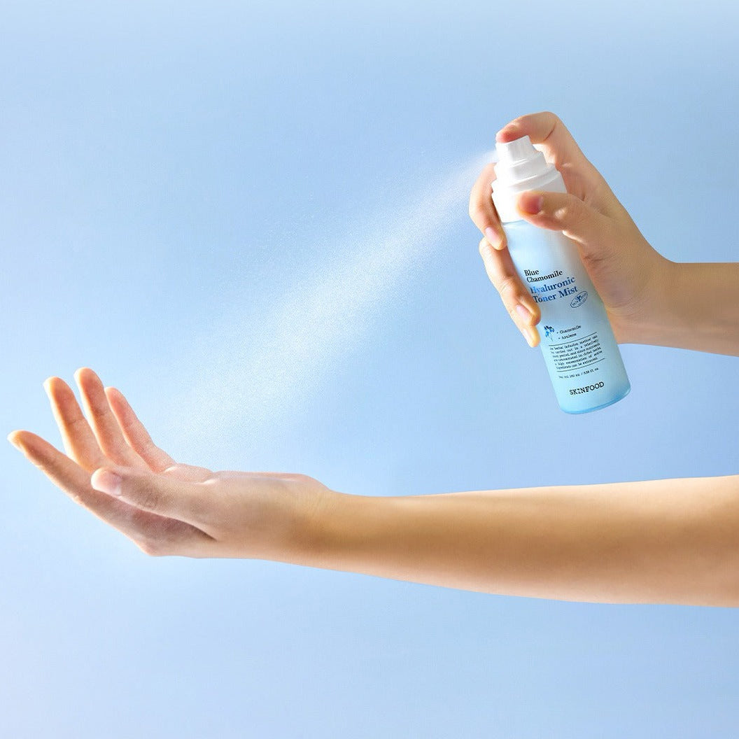 Person spraying SKINFOOD Blue Chamomile Hyaluronic Toner Mist 100ml onto hand against blue background.