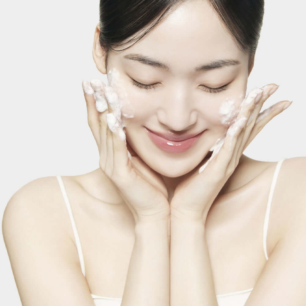 Woman using SU:M37 Secret Rice Nuruk Peeling to Foam Cleanser 120g with gentle bubbles on cheeks, showcasing skincare routine.