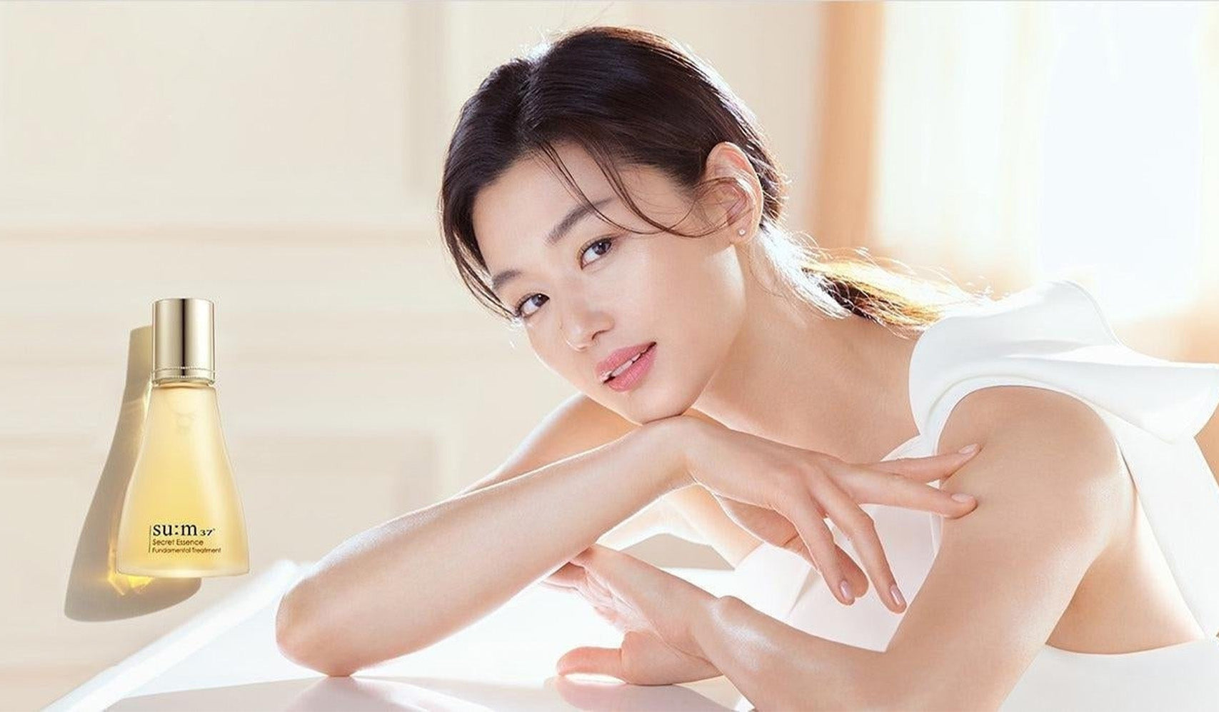 Woman posing with Sumi skincare bottle, radiant and glowing complexion, in soft light setting.