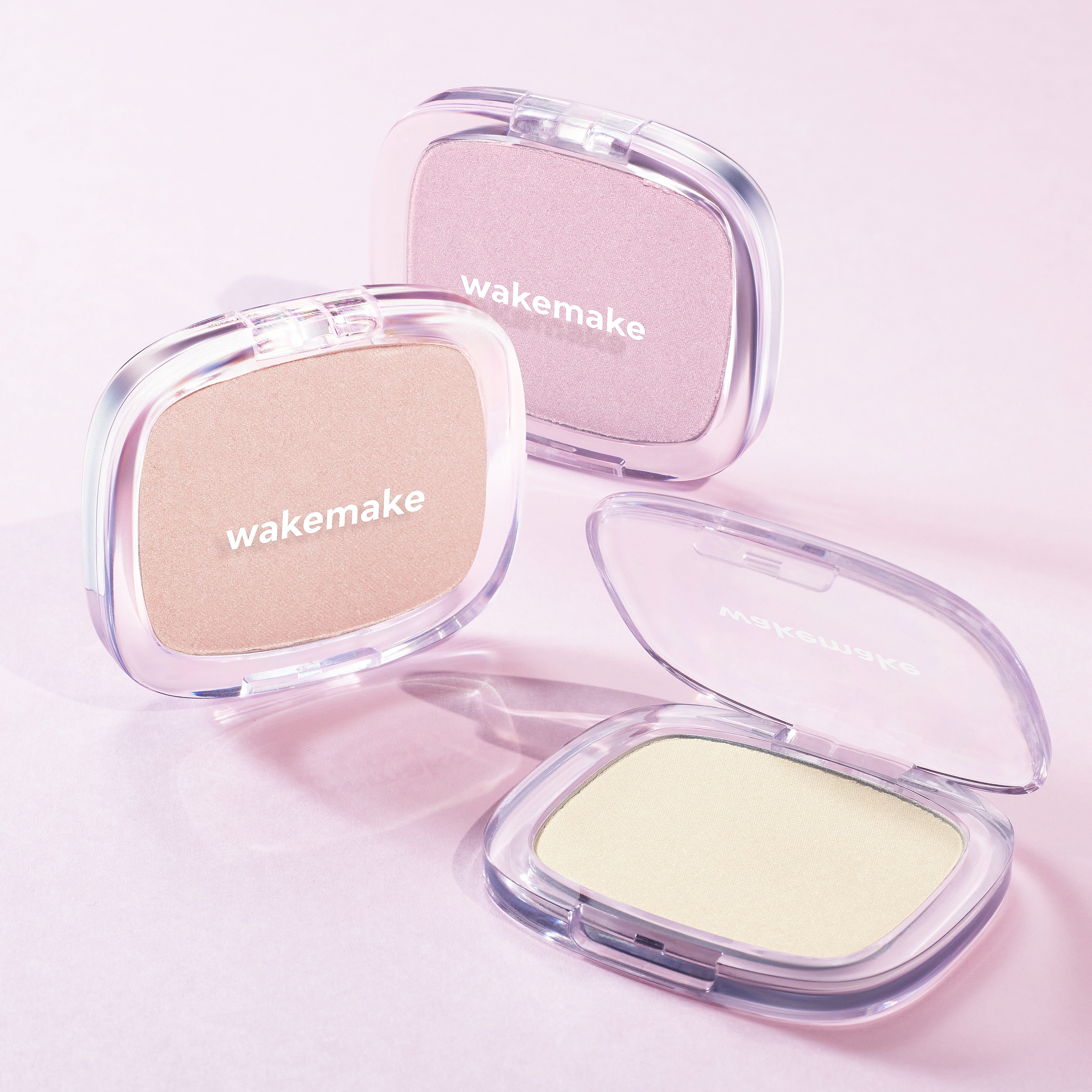Three WAKE MAKE Sheer Breeze Highlighter 5g compacts in different shades on a pink background.
