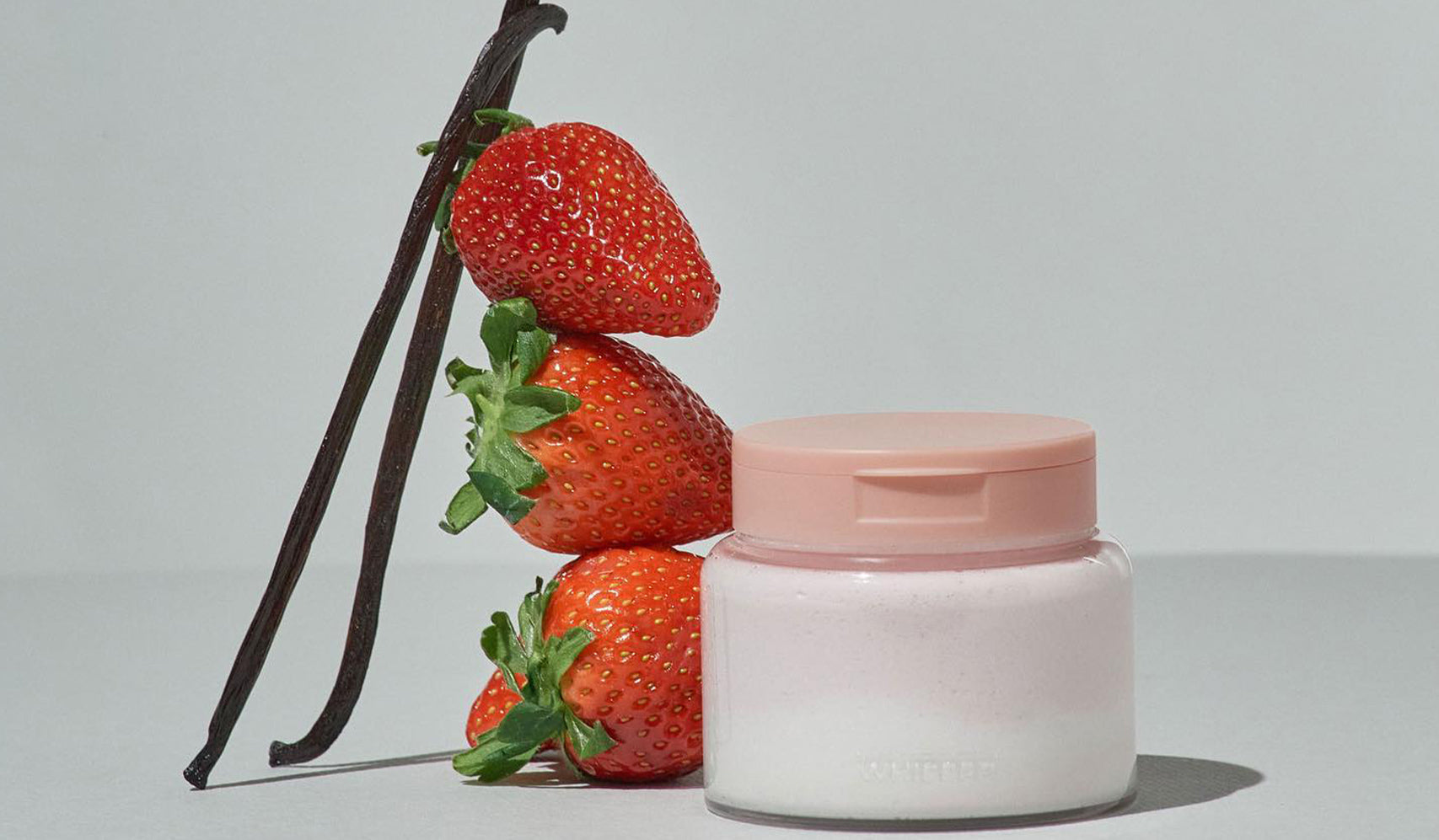 Vanilla and strawberry skincare cream in jar, next to fresh strawberries and vanilla beans, on a minimalist background.