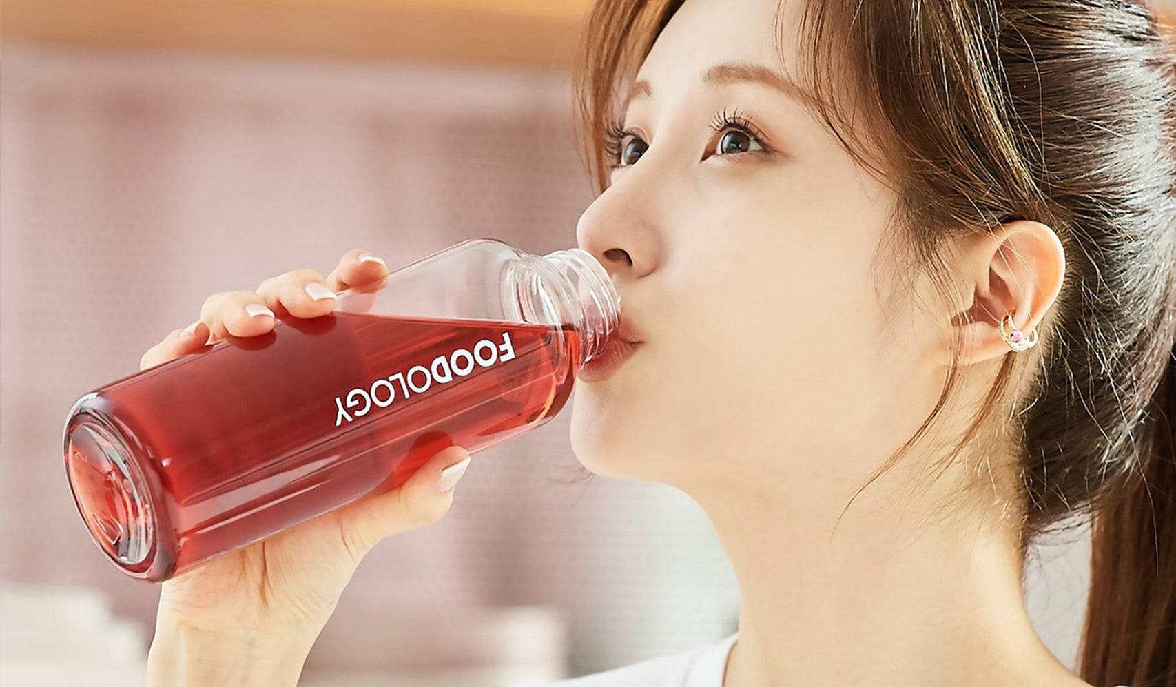 Person drinking red beverage from a glass bottle marked Foodology indoors