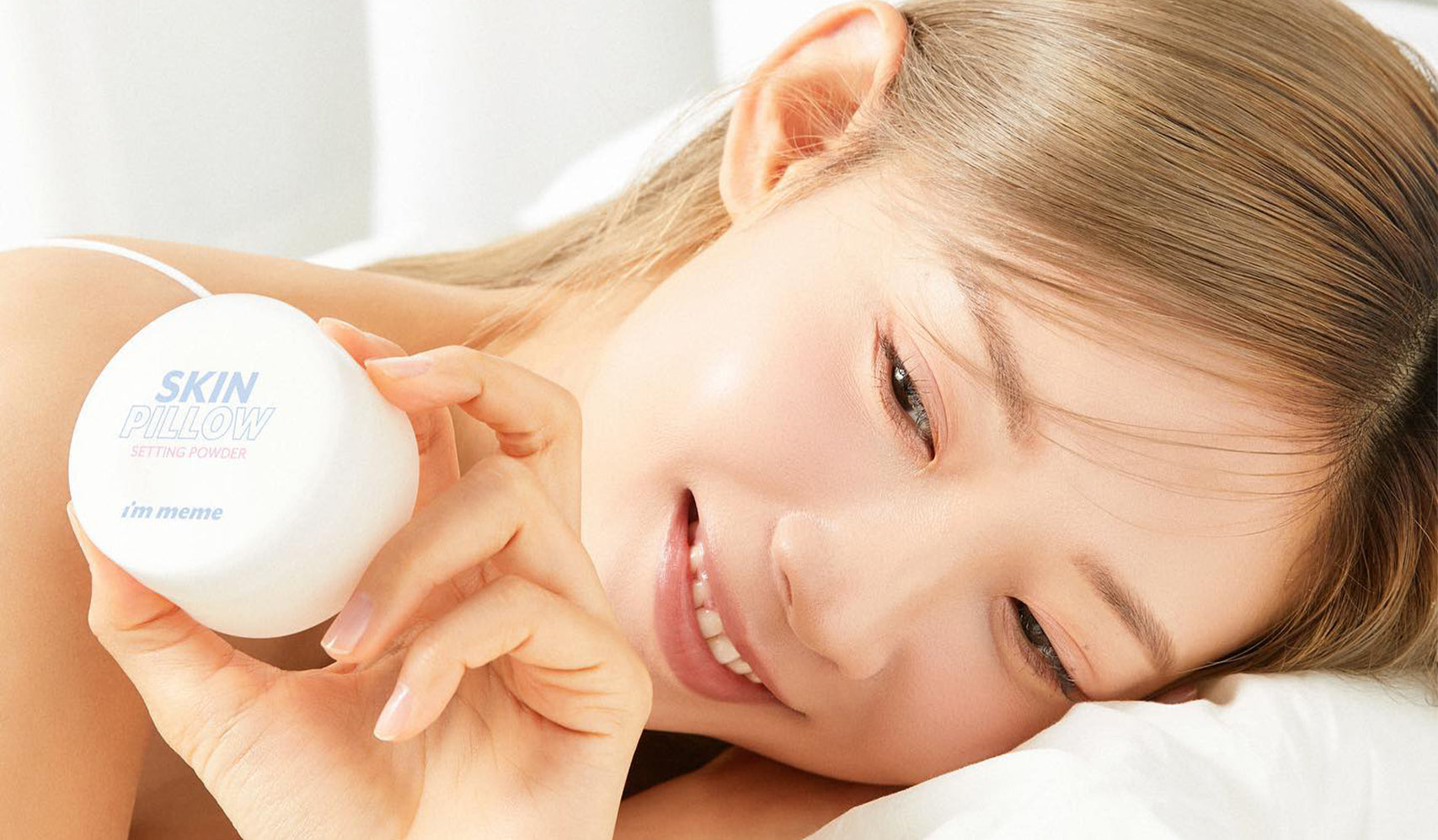 Person holding a skincare product container labeled 'Skin' while smiling, lying on a white bed, highlighting a skincare routine moment.