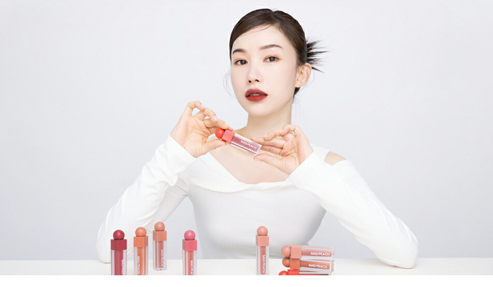 Woman showcasing lip gloss collection in various shades on a table, wearing white top and red lipstick.