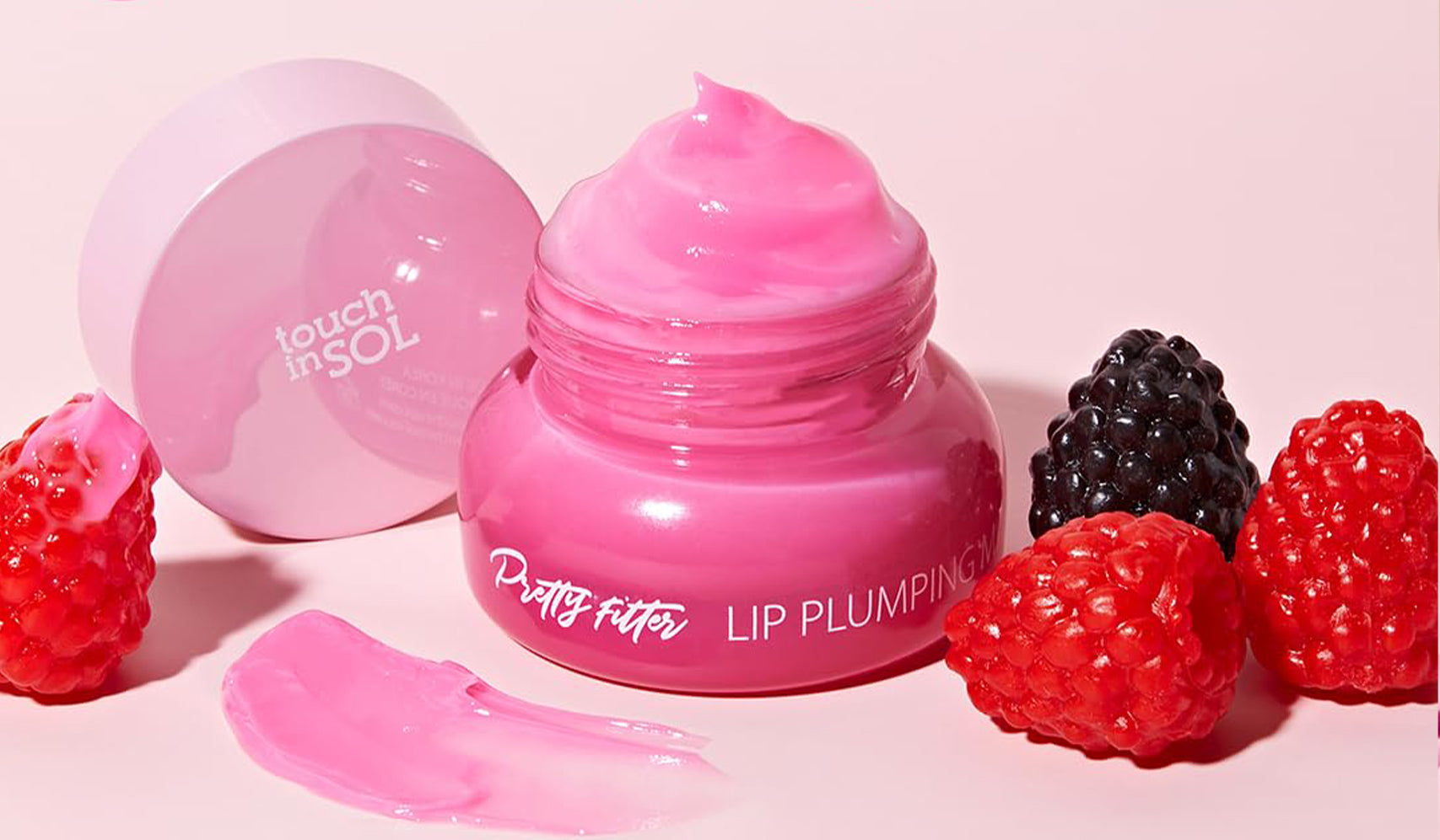 Pink lip plumping balm in open jar surrounded by raspberries and blackberries on a pink background.