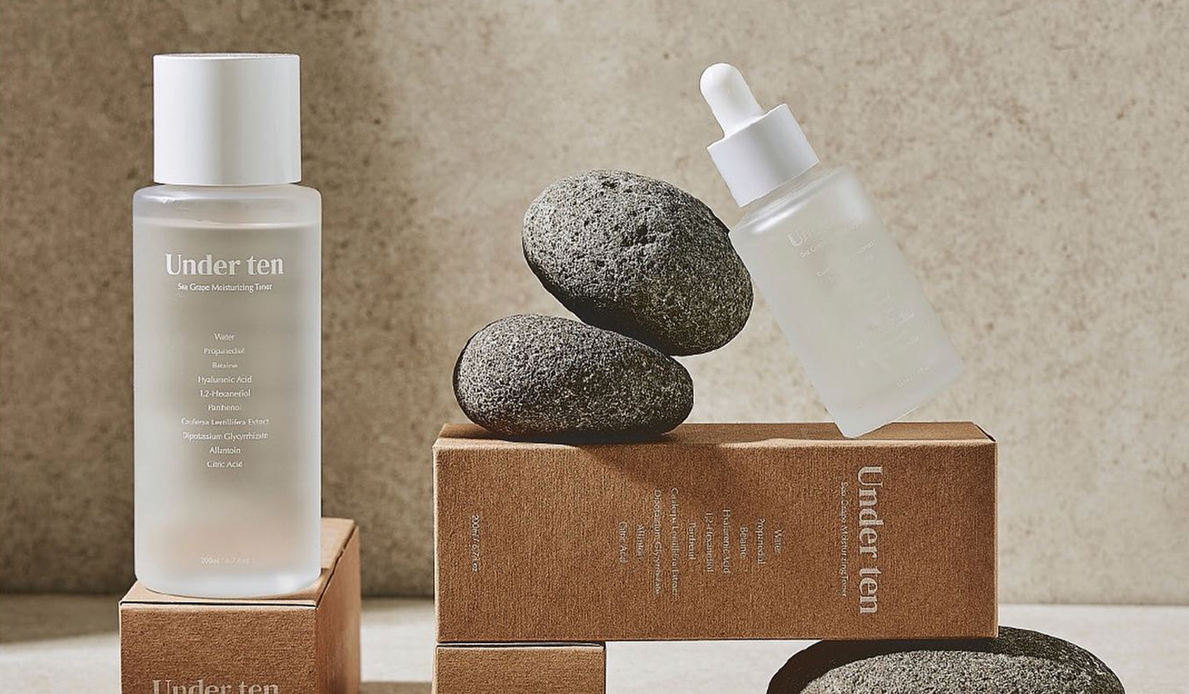 Two skincare bottles labeled 'Under Ten' with brown packaging, displayed elegantly on and around smooth gray stones.