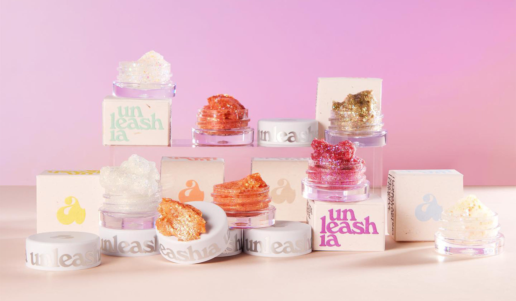 Colorful jars of cosmetic glitter on a pink background, featuring Unleashia packaging and shimmering products on display.